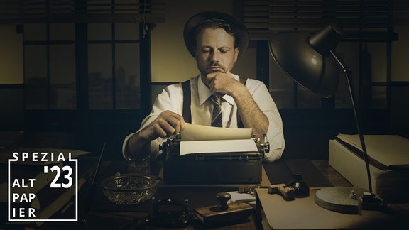Ein Mann sitzt vor einer Schreibmaschine und blickt kritisch auf das bisher Geschriebene.