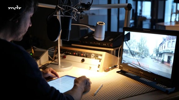Ein Mann sitzt in einem Tonstudio vor einem Mikrophon. Schräg vor ihm auf dem Tisch seht ein Monitor auf dem ein Filmausschnitt zu sehen ist. 