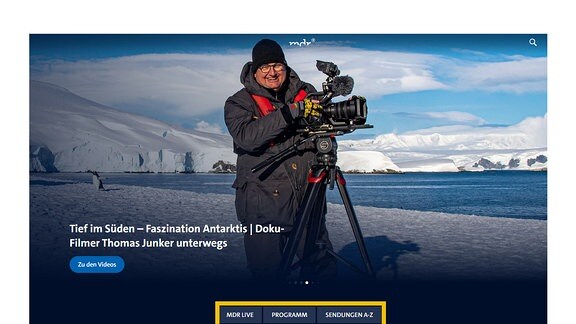 Das Bild zeigt einen Screenshot der MDR-Mediathek. Ein Vorschaubild einer Sendung ist zu sehen. Es zeigt den Thomas Junker mit einer Kamera auf einem Stativ. Im Hintergrund ist eine verschneite Berglandschaft und eine Uferbereich zu sehen. Dahinter das Meer. Das Bild trägt die Überschrift: "Tief im Süden - Faszination Antarktis: Doku-Filmer Thomas Junker unterwegs. Darunter befindet sich die Schaltfläche "Zu den Videos". Am unteren Bildschirmrand befinden sich 3 weitere Schaltflächen. Von links nach rechts:"MDR LIVE", "PROGRAMM" und "SENDUNGEN VON A-Z.