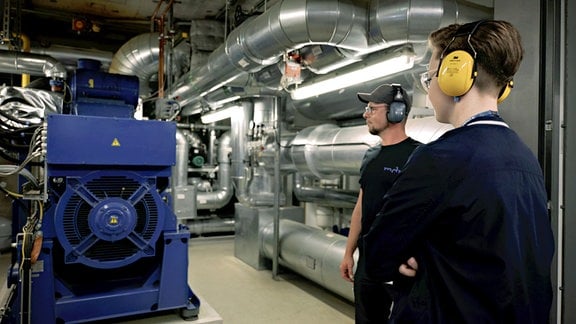 Zwei junge Menschen stehen mit Ohrenschutz vor einem blauen Maschinengehäuse. Im Hintergrund sind große silberne Leitungen zu sehen.