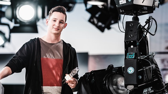 Junger Mann steht in einem Fernsehstudio neben einer Kamera.