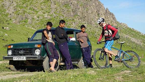 Drei Männer lehnen an einem Auto, daneben ein Mountainbiker