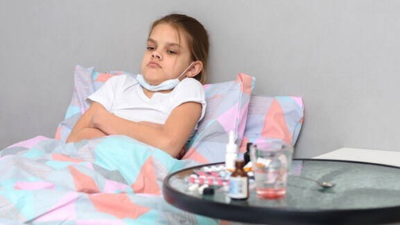 Ein Kind mit runtergezogenem Mundschutz liegt im Bett