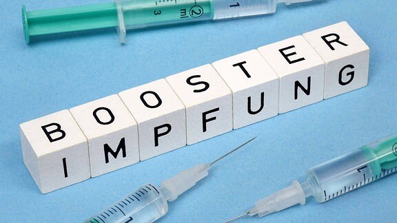 Neben drei Spritzen befinden sich Buchstabenwürfel mit dem Schriftzug Boosterimpfung.