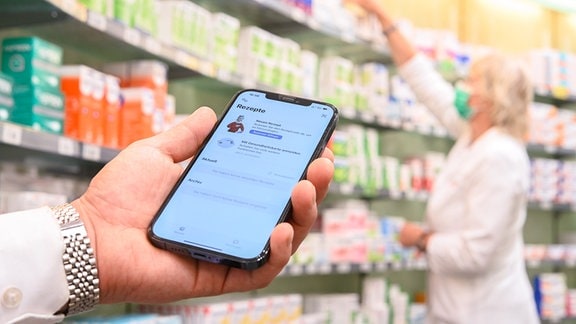 Auf einem Smartphone ist die geöffnete App «Das E-Rezept» zu sehen, während im Hintergrund eine Apothekerin in einer Apotheke an einem Regal steht. 