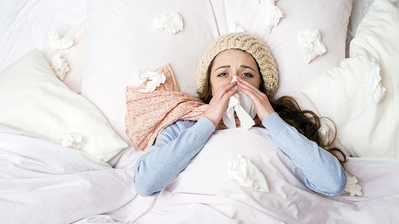 Eine erkältete Frau liegt schniefend und triefend im Bett.