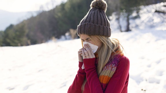 Junge blonde Frau mit Mütze steht in einer Winterlandschaft und putzt sich die Nase.