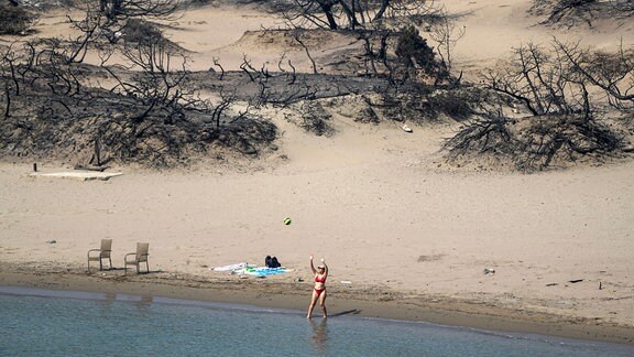 Eine Frau spielt mit einem Ball vor einem verbrannten Wald an einem Strand in der Nähe des Dorfes Gennadi.