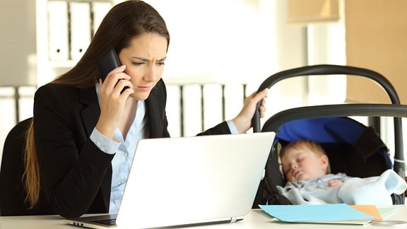 Symbolbild Stress im Homeoffice - Mutter, das Kind schaukelnd und telefonierend am Laptop