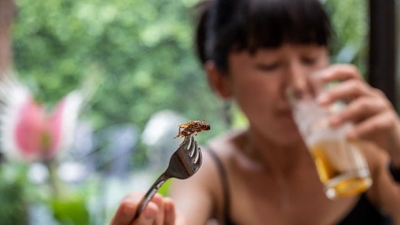 Eine Frau mit einem Insekt auf einer Gabel trinkt aus einem Glas.