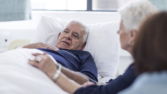Ein älterer Mann liegt in einem Krankenhausbett, daneben sitzen Angehörige