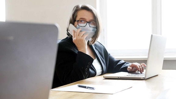 Frau im Büro am Arbeitsplatz mit Mundschutz um Ansteckung bei Kollegen zu vermeiden