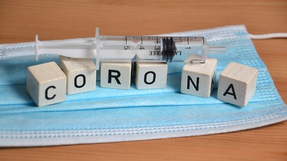 Auf einem medizinischen Maske bilden Buchstabenwürfel den Schriftzug 'CORONA', darüber liegt eine Spritze.