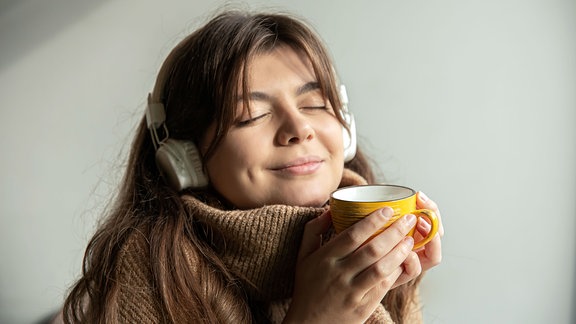 Junge Frau in einem gemütlichen Pullover hört mit einer Tasse in der Hand Musik über Kopfhörer.