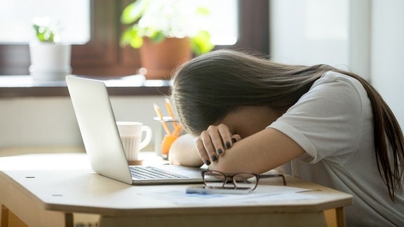 Eine Frau hat den Kopf auf den Armen liegend vor einem Laptop