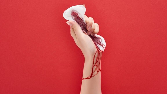 Eine Hand hält eine Monatsbinde vor rotem Hintergrund.
