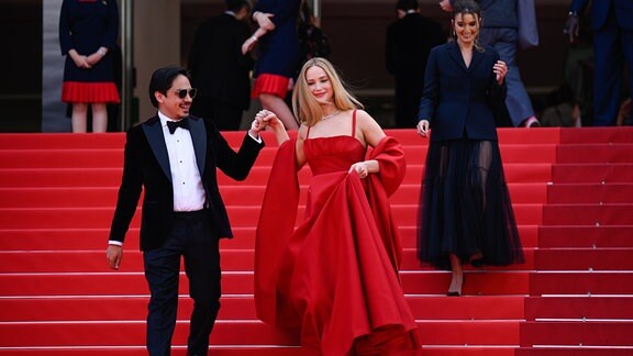 Eine junge Frau in rotem Abendkleid und ihre Begleitung gehen eine Treppe hinunter.