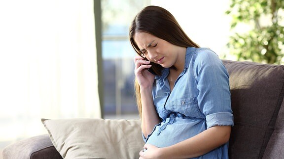 Eine schwangere Frau, die sich schmerzerfüllt den Bauch hält, telefoniert. 