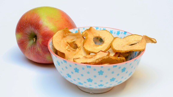 Ein Apfel liegt hinter einer Schale mit getrockneten Apfelringen.