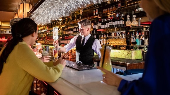 Frauen sitzen an einer Bar, ein Barkeeper mixt ein Getränk. 
