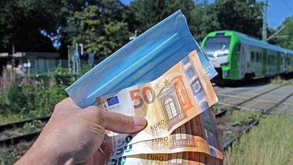Maske und Geldscheine werden in einer Hand gehalten, im Hintergrund eine grüne Regionalbahn.