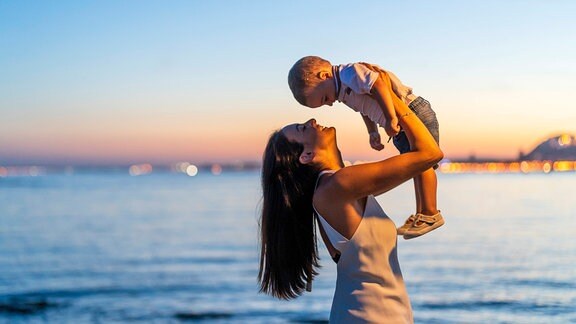 Frau hebt Kind an Strand bei Sonnenuntergang