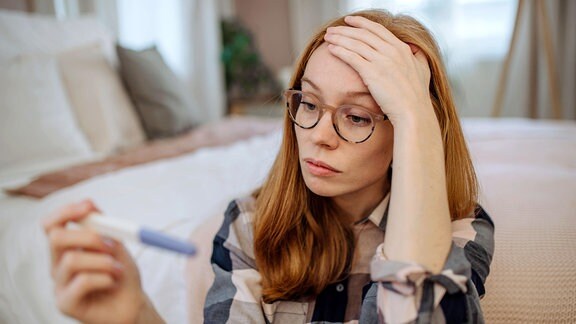 Besorgte Frau mit Brille sitzt mit einem Schwangerschaftstest-Kit vor dem Bett zu Hause.