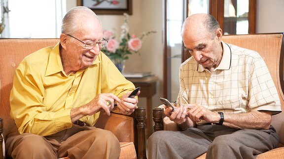 Zwei Senioren sitzen in Sesseln und sehen auf ihre Telefone