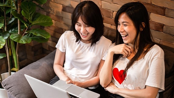 Zwei junge Frauen sitzen lachend auf einer Couch am Laptop