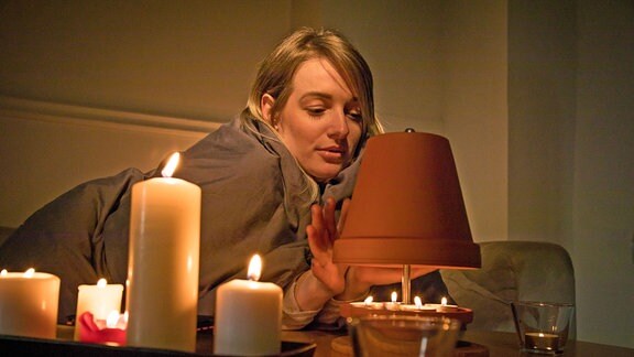 Eine junge Frau sitzt, umgeben von Kerzen in einer Wohnung auf Grund eines Stromausfalls im Dunkeln und wärmt sich an einer Teelichtheizung