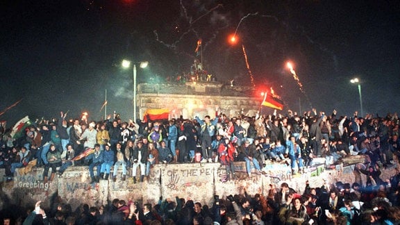 Zahlreiche Menschen sitzen auf und stehen vor der Berliner Mauer, im Hintergrund ist das Brandenburger Tor zu sehen, Pyrotechnik leuchtet in der Luft.