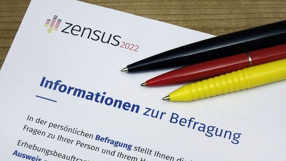 Auf einem Informationsblatt zum Zensus 2022 liegen Kugelschreiber in den Deutschlandfarben