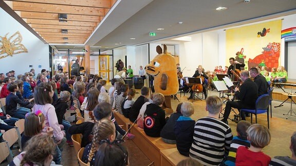 Die Maus aus der "Sendung mit der Maus" steht mit Schulkindern und MDR-Musiker*innen auf der Bühne eines MDR-Clara-Schulkonzerts in Radeburg.