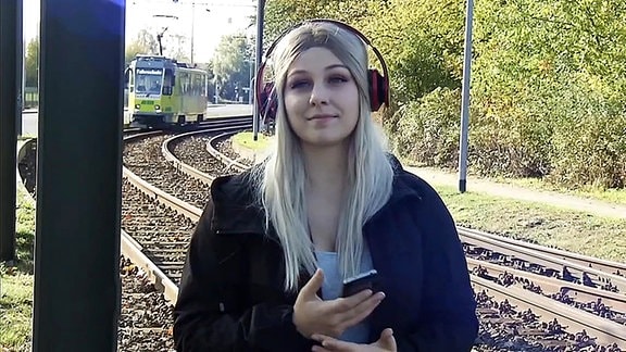 Eine junge Frau mit Kopfhörern, hinter ihr fährt eine Straßenbahn.