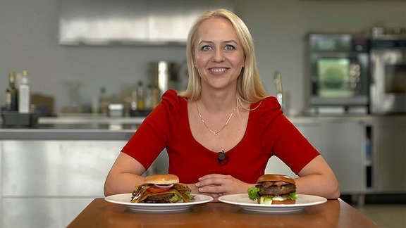Die Moderatorin sitzt vor zwei Tellern, auf denen je ein Burger liegt.