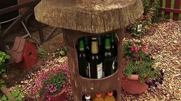 In einem Garten steht ein aus dem Boden fahrbarer Holzschrank mit Bierflaschen im Innern.