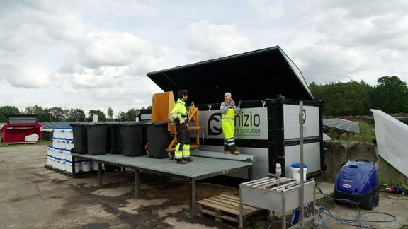 Zwei Personen in Schwarzgelber Arbeitsschutzkleidung stehen auf einem Podest vor einem speziellen Container.