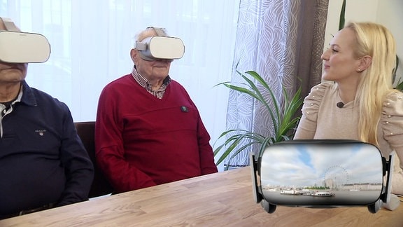 Zwei ältere Herren sitzen mit einer VirtualReality Brille an einem Tisch, daneben eine junge blonde Frau die lächelnd schaut.