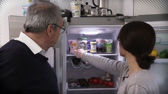 Zwei Personen stehen vor einem geöffnetem Kühlschrank. Eine Frau greift nach etwas.