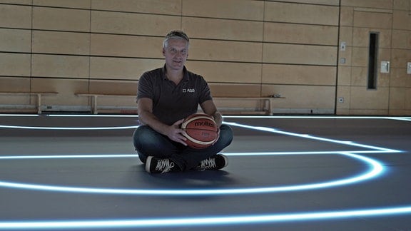 Ein Mann sitzt mit einem Basketball auf einem Sporthallenboden.