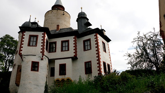 Blick auf eine verschachtelt wirkende Burg mit mehreren Türmen, die auf einem begrünten Hügel thront. 