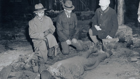 Mehrere Personen knien vor einer Leiche in einer Scheune.