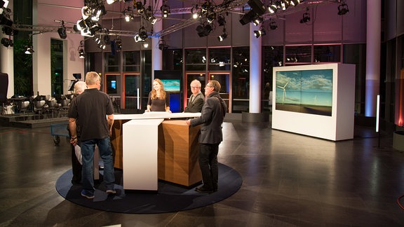 In einem Fernsehstudio stehen eine Frau und fünf Männer im Kreis um einen Tisch.