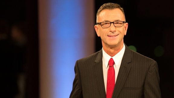 Ein dunkelhaariger Mann in Anzug und roter Krawatte und mit Brille blickt in eine Kamera und lächelt.