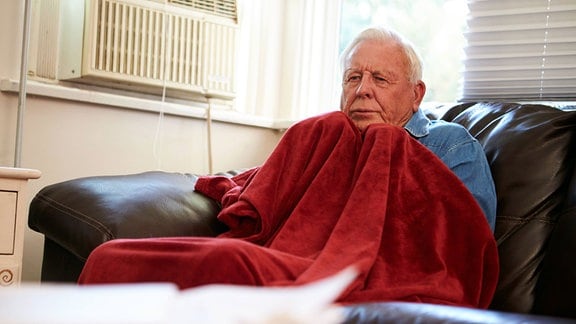 Ein Senion sitzt frierend, in eine Decke gehüllt auf dem Sofa.