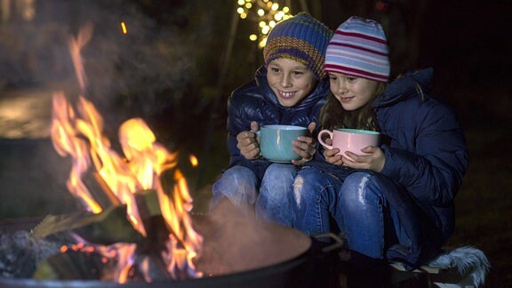 Junge und Mädchen sitzen mit Bechern in den Händen an einem Feuer.