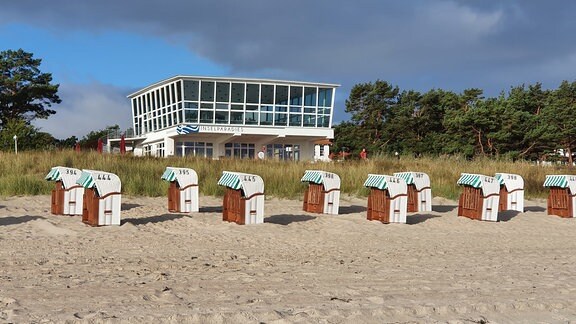 Weißer Sandstrand mit Strandkörben, im Hintergrund ein Gebäude mit großer Glasfassade