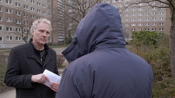 Zwei Männer stehen im Gespräch vertieft vor einem Hochhaus. Um nicht erkannt zu werden, wurde ein Mann mit Kaputze von hinten gefilmt.