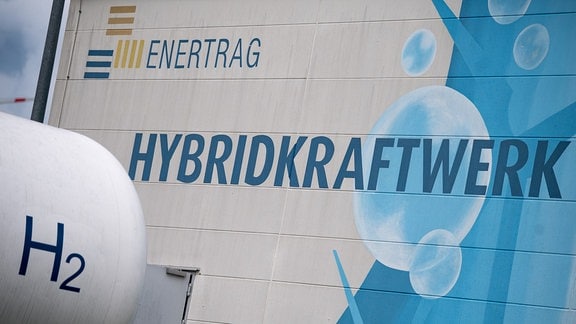 Ein Wasserstoffspeicher steht auf dem Gelände des Hybridkraftwerks Enertrag, während im Hintergrund auf einem Gebäude der Schriftzug "Hybridkraftwerk" zu sehen ist