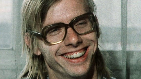 Gerhard Gundermann: ein junger Mann mit langen blonden Haaren und einer Brille mit breitem Rand lacht.